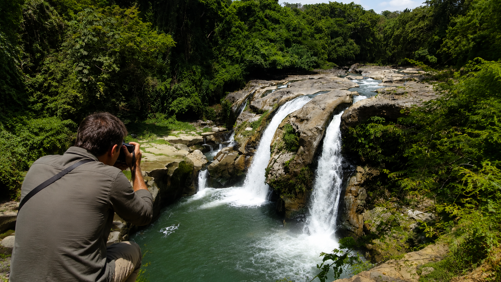 A photographer crouching with his camera pointed at Salto de Malacatiupán, a turquoise waterfall surrounded by tropical forest in El Salvador