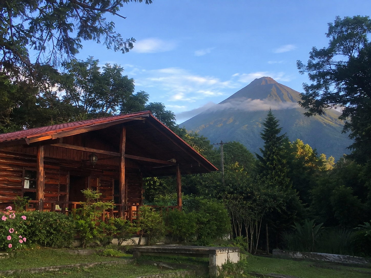 A rustic wooden cabin with a red roof nestled in a lush green garden with Izalco Volcano rising in the background, El Salvador.